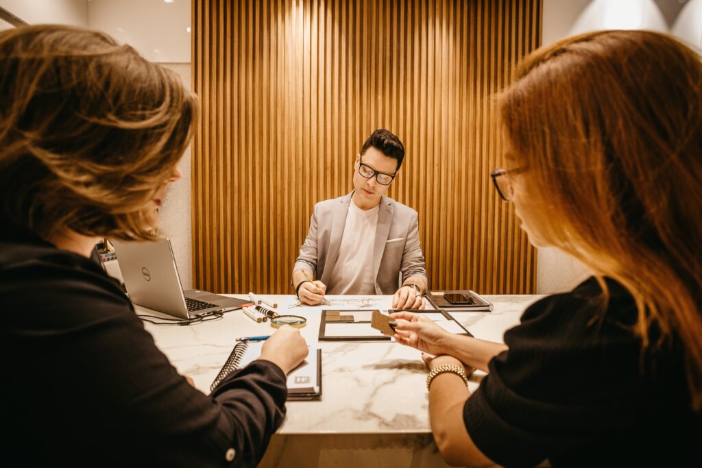 Man in a gray blazer sits at a table in front of two women in meeting.