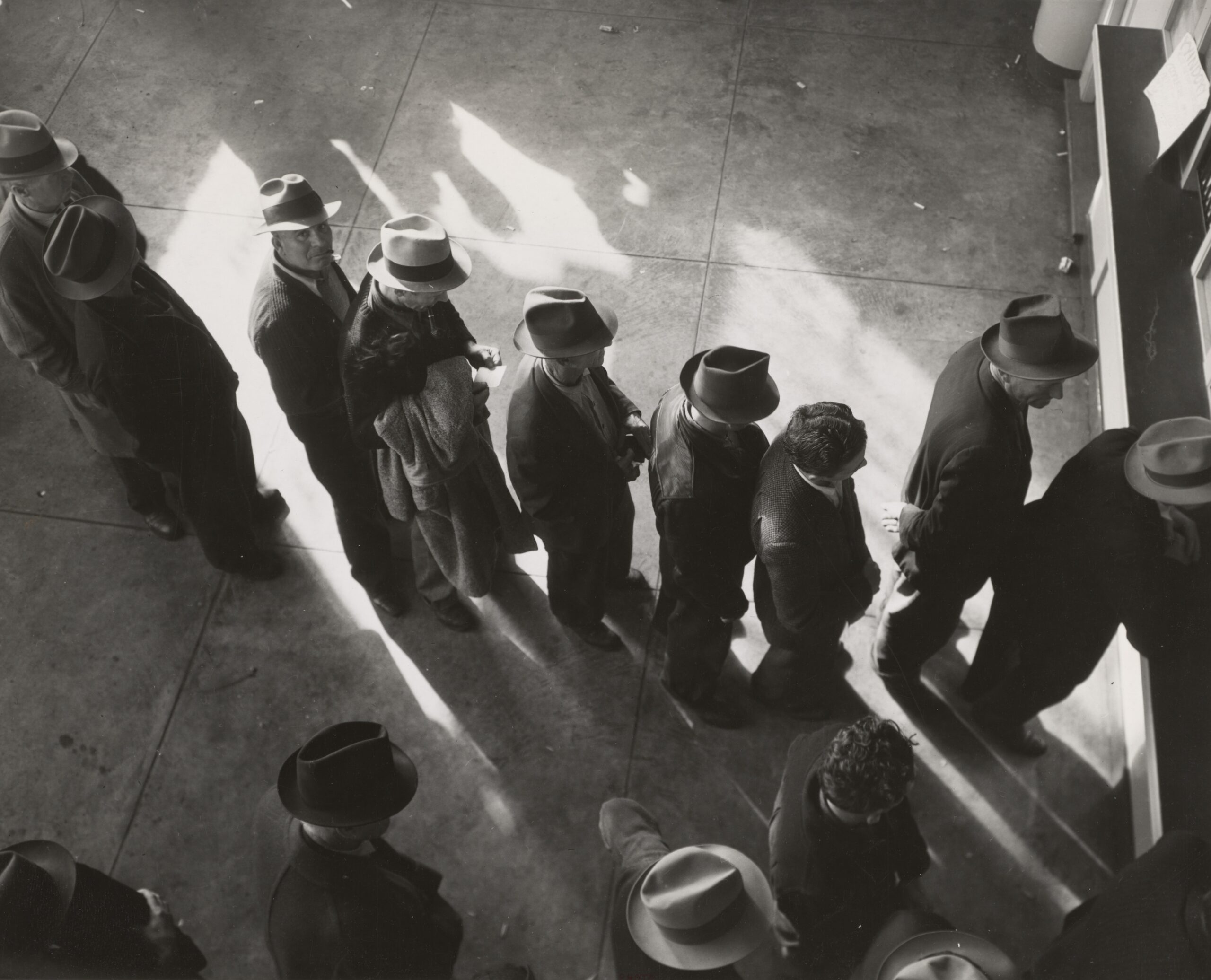 1938 - Unemployment benefits aid begins. Line of men inside a division office of the State Employment Service office at San Francisco, California, waiting to register for benefits on one of the first days the office was open. Photographer - Dorothea Lange