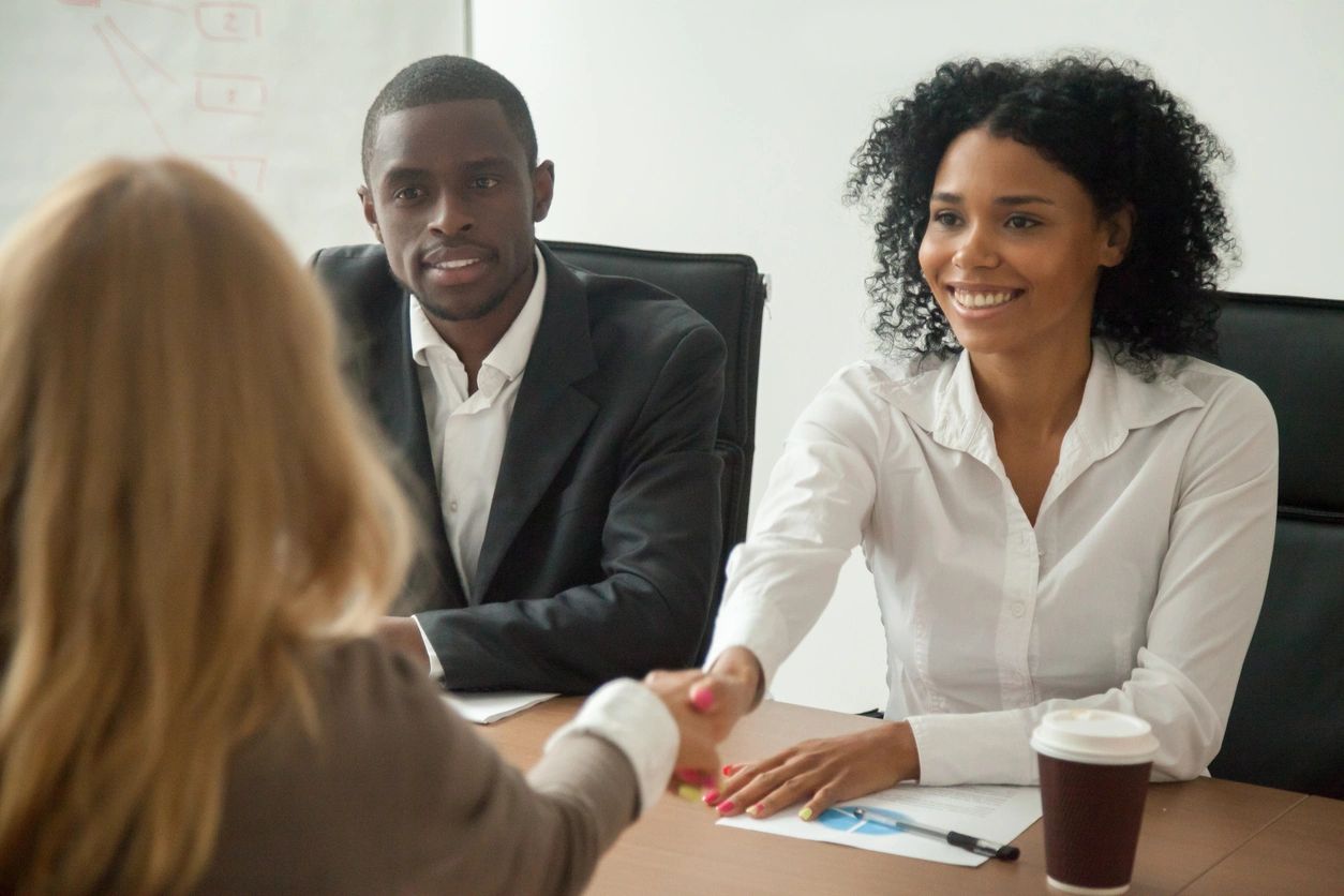 Two business women shake hands next to a business man over a conference table.